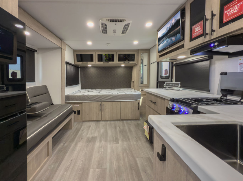 Interior of a modern RV featuring a compact open layout with a small kitchen, black sink, stovetop, oven, and overhead microwave on the right. A queen bed with storage drawers and overhead cabinets is centered at the back, while a cushioned bench sits to the left. The space has light wood cabinetry, gray flooring, and recessed ceiling lights, creating a bright and contemporary look.