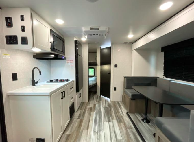 Interior of a modern travel trailer featuring a compact kitchen with white cabinetry, a black faucet, stovetop, microwave, and overhead storage on the left. The right side has a booth-style dinette with a dark tabletop and cushioned seating under a large window with a black blind. The space has light wood-look flooring, recessed ceiling lights, and leads to a rear sleeping area with visible bunks through an open doorway.