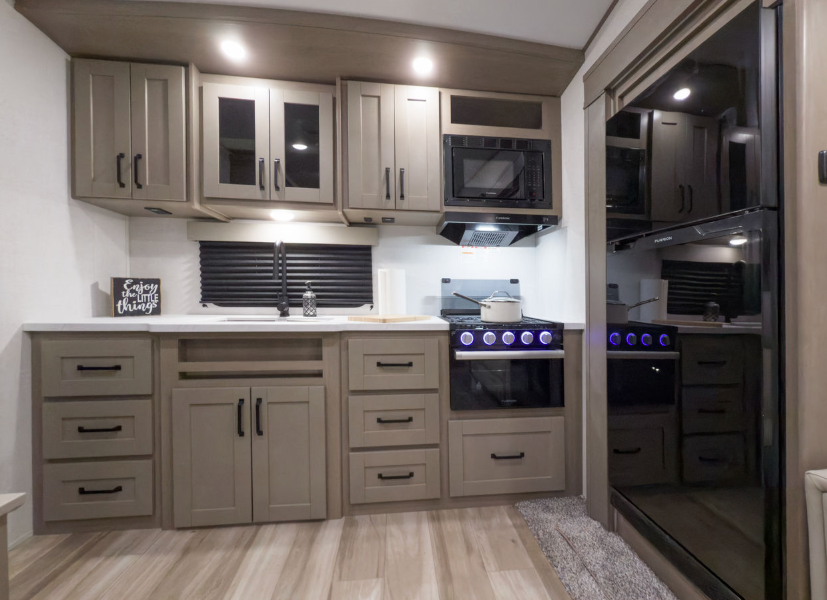 Interior view of an RV kitchen featuring modern cabinetry in a muted tan finish with black hardware, a stainless steel microwave above a stovetop with blue-lit knobs, and ample counter space. Multiple drawers and cabinets provide storage, while a full-size refrigerator is visible on the right. The flooring is light wood-look vinyl, and under-cabinet lighting brightens the space.
