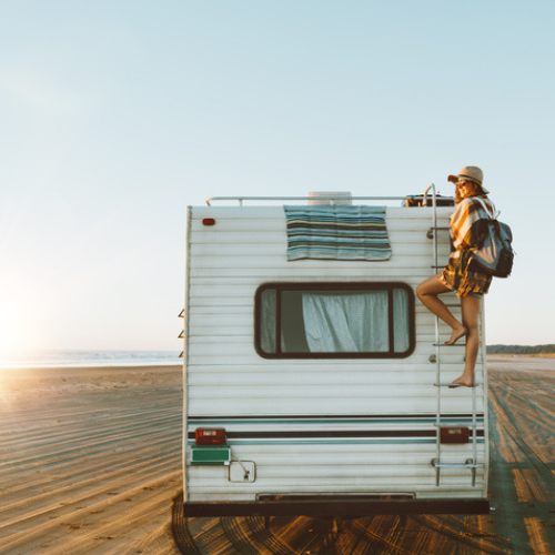 A person in a sunhat and backpack climbs the rear ladder of a white RV parked on a sandy beach at sunset, with a towel draped over the back window and the ocean visible in the distance.