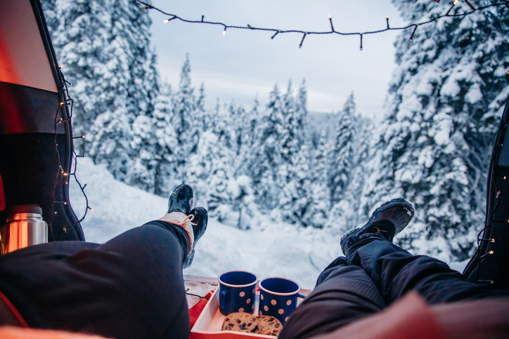 View from inside a tent looking out over a snowy winter forest. Two people’s legs in snow boots are stretched out toward the scene, with a tray holding two blue mugs and cookies between them. String lights hang across the top of the tent opening, adding a cozy touch to the snowy landscape.