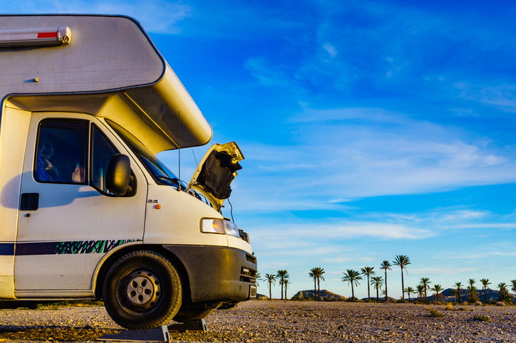 An RV with its hood open parked in a desert setting with palm trees in the background, under a bright blue sky with scattered clouds, suggesting the RV has broken down during travel.