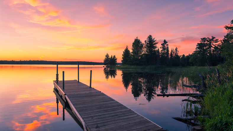 A peaceful lakeside scene at sunset with a wooden dock extending into calm, reflective water. The sky glows with warm hues of orange, pink, and purple, mirrored in the lake’s surface, while tall trees line the shoreline on the right.