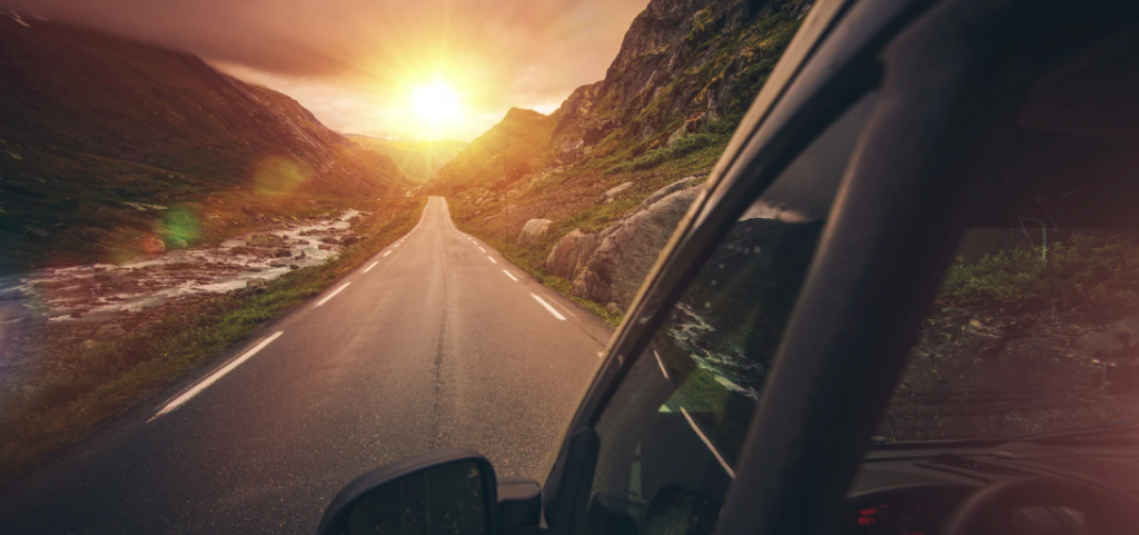 Scenic road trip view from an RV window, showing a winding mountain road at sunset with a glowing sun over rocky cliffs and a flowing river alongside the road.