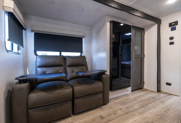 Modern RV interior featuring a dark gray sectional sofa with tufted cushions. A cream-colored throw blanket is draped over the backrest, and a small detachable table with a cupholder is attached to one seat. A large window lets in natural light, with a dining area partially visible in the background, creating a cozy and functional living space.