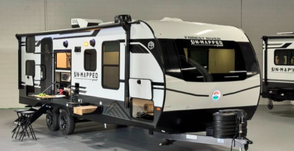 Forest River R-Pod Unmapped travel trailer with white and black exterior, outdoor kitchen setup, and modern design, displayed indoors on a showroom floor.