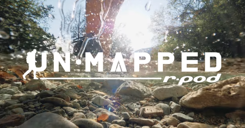 A close-up of a hiker’s feet splashing through a shallow rocky stream with water droplets frozen mid-air. The sun shines brightly through the trees in the background, creating a vibrant, adventurous atmosphere. Overlaid on the image is the logo text “UN•MAPPED r.pod” with a small hiker icon.
