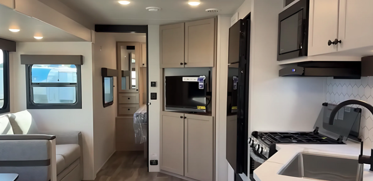 Interior of a modern travel trailer kitchen with light cabinetry, microwave, gas stove, stainless sink, and view toward the hallway and seating area.