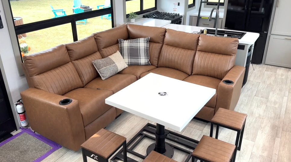 Spacious RV living area in the Axiom Imperium 12900 featuring an L-shaped brown leather reclining sofa with cupholders, dining table with stools, and modern kitchen with stainless steel appliances.