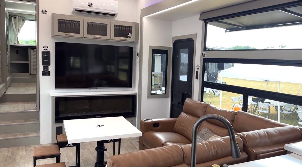 Luxury RV living room in the Axiom Imperium 12900 featuring a reclining leather sofa, entertainment center with flat-screen TV and fireplace, and panoramic windows for natural light.