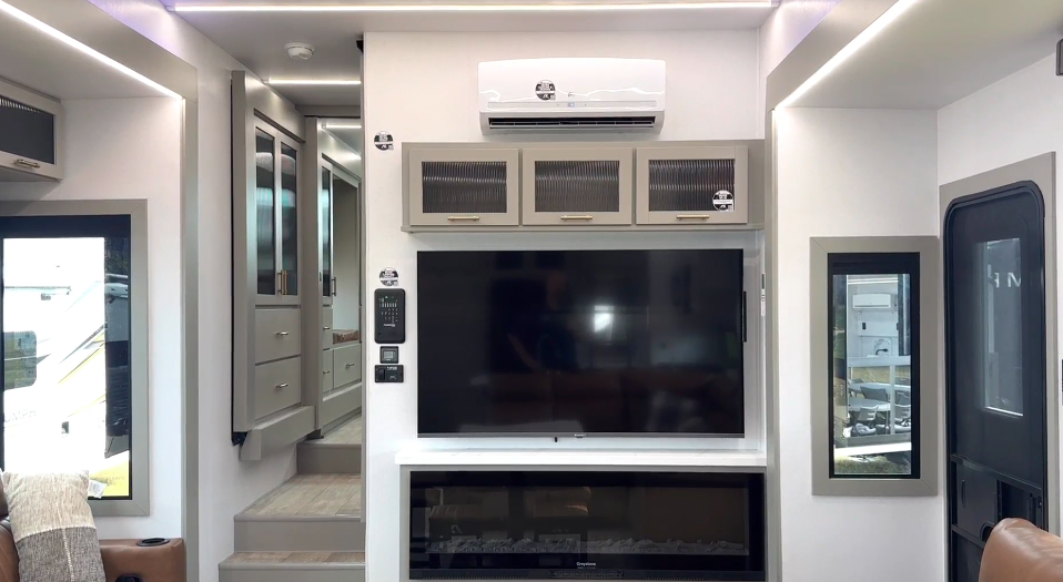 Modern RV entertainment center in the Axiom Imperium 12900 featuring a large flat-screen TV, electric fireplace, overhead storage cabinets, and energy-efficient mini split air conditioning system.