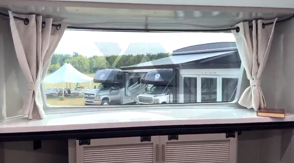 Large panoramic bedroom window with curtains inside the Axiom Imperium 12900 fifth wheel RV, bringing in natural light above built-in storage cabinets.