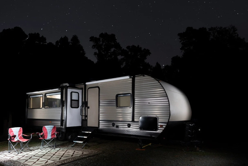 Travel trailer parked at a campsite under a starry night sky with outdoor lights illuminating two red camping chairs and a rug, creating a cozy RV camping setup.