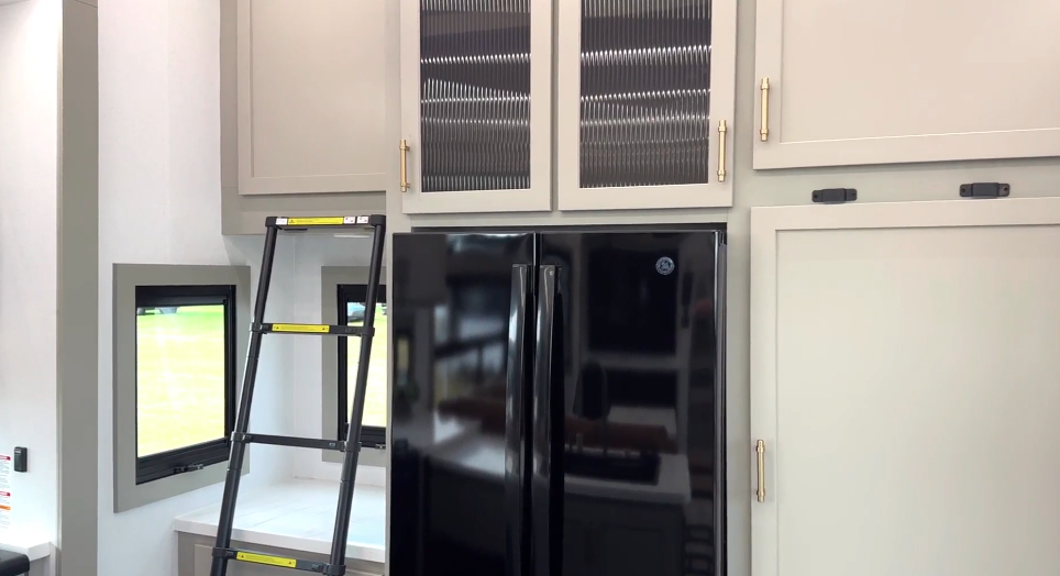 Residential refrigerator with surrounding cabinetry and built-in ladder access to upper storage inside the Axiom Imperium 12900 RV kitchen.
