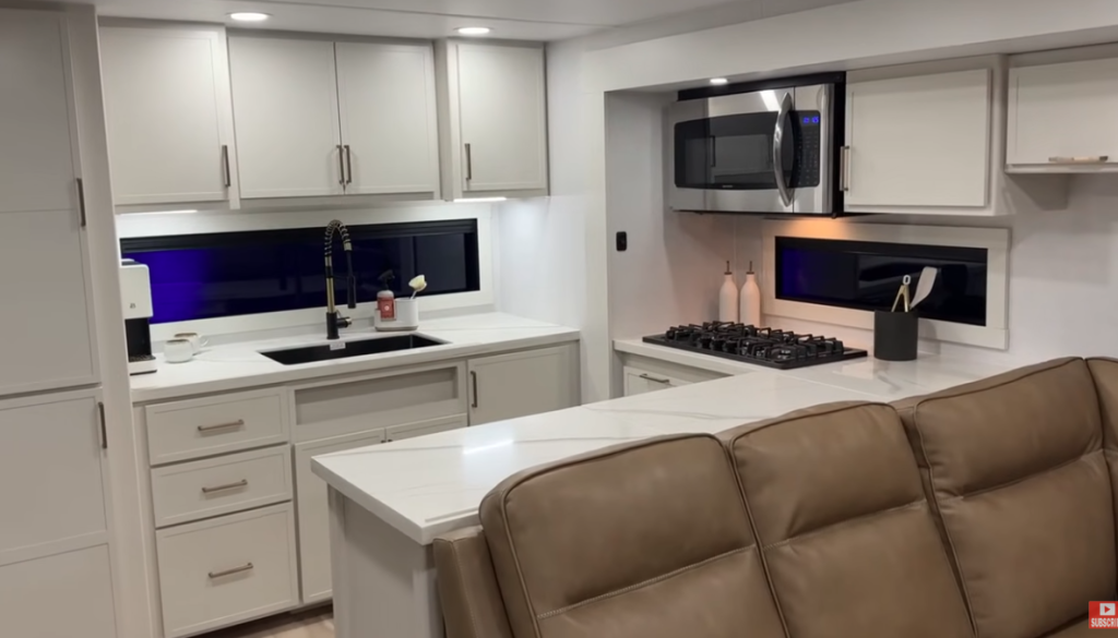 Brinkley RV kitchen with stainless steel microwave, gas cooktop, farmhouse sink, white cabinetry, and modern backsplash beside leather seating.