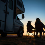 A couple sits in camping chairs beside their motorhome at sunset. The RV is parked on a beach or open area, and the two people face each other in conversation as the sun sets on the horizon, creating a warm glow and long shadows.