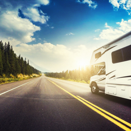 A white motorhome drives down an empty two-lane highway surrounded by tall pine trees, with the sun shining brightly in a partly cloudy blue sky, creating a sense of freedom and adventure on the open road.