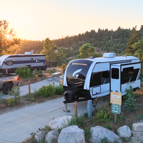 A white Wayfinder travel trailer parked at a campsite during sunset, with a pickup truck and another RV nearby. The trailer is set up on a concrete pad with surrounding greenery, rocks, and a sign marking campsite numbers. The warm golden light highlights the RV and trees in the background.