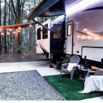 Modern RV parked at a wooded campsite with string lights and an extended awning, featuring cozy outdoor seating with chairs, blankets, and a small table on artificial grass.