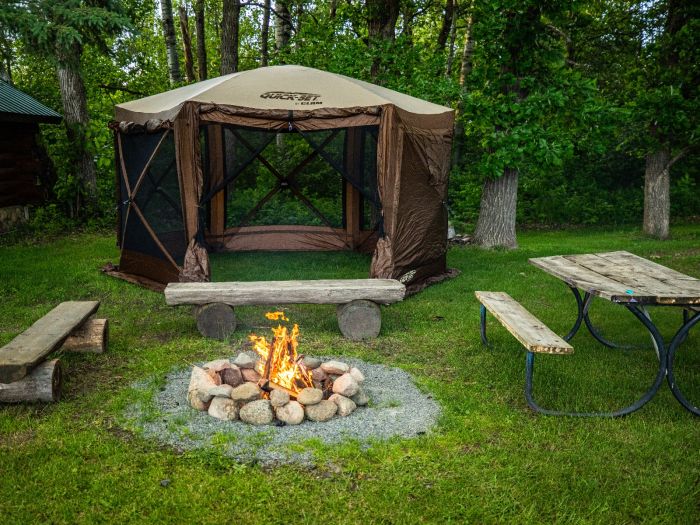Campsite setup with a screened brown pop-up canopy tent, surrounded by wooden benches and a picnic table, featuring a lit stone campfire in the grassy clearing
