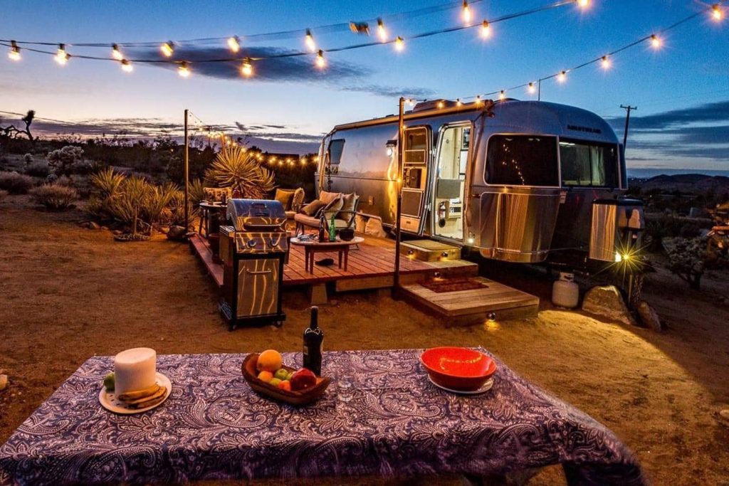 Luxury Airstream travel trailer parked at desert campsite with string lights, outdoor deck, grill, and cozy lounge setup at sunset