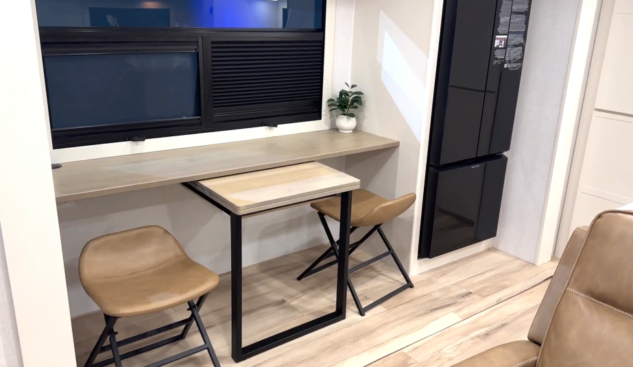 Multifunction dinette and workspace inside the Brinkley Model I 280, featuring a sleek fold-out table with two tan leather chairs and a built-in counter under large dual-pane windows. A black 12V refrigerator stands nearby, and a small potted plant adds a touch of home comfort, highlighting the RV’s modern and versatile design.