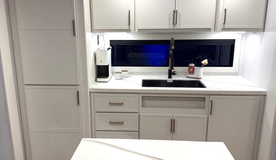 Modern RV kitchen sink area inside the Brinkley Model I 280 featuring solid-surface countertops, matte black faucet, and white cabinetry with brushed metal handles. A compact coffee maker, mugs, and cleaning accessories sit beside the large horizontal window, creating a bright, residential-style workspace with under-cabinet lighting.