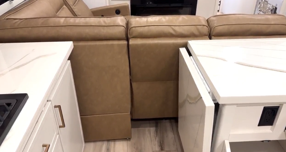 Close-up view of the Brinkley Model I 280 kitchen island showing the solid-surface waterfall countertop extension with power outlet and soft-close drawer. Behind it, a tan leather quad-recliner faces the entertainment center, highlighting the RV’s seamless blend of residential comfort and smart, functional design.