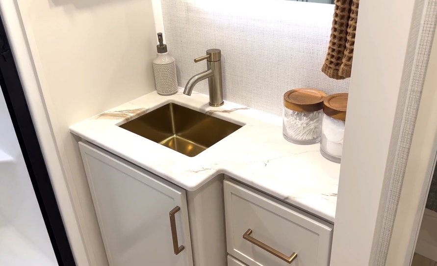Bathroom vanity inside the Brinkley Model I 280 featuring a solid-surface countertop with marble-style finish, undermount sink with brushed gold faucet, and soft-close cabinetry. The setup includes decorative jars and a textured hand towel, creating a modern, spa-like feel with practical storage and refined residential design.