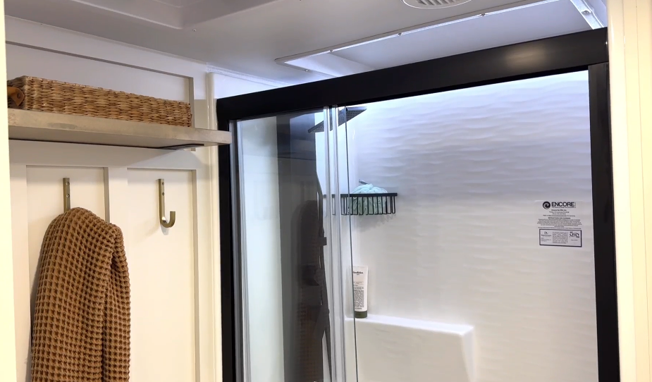 Bathroom shower area inside the Brinkley Model I 280 featuring a one-piece fiberglass shower with textured white walls, black-framed sliding glass doors, and a built-in seat. Overhead recessed lighting and a nearby robe hook with shelf storage create a bright, spa-like space designed for durability and comfort.