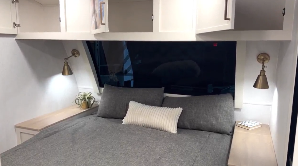 Bedroom inside the Brinkley Model I 280 featuring an Olympic queen bed with gray bedding, overhead storage cabinets, brass reading lamps, and dual nightstands. A large front windshield window adds natural light and openness, creating a bright, modern, and residential feel within the RV’s private sleeping space.