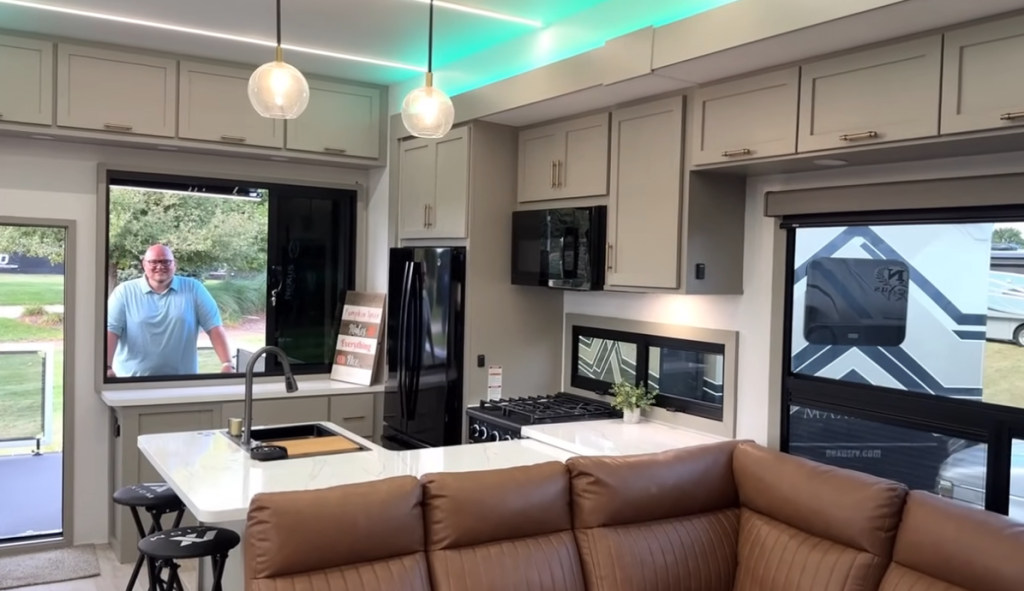 Modern kitchen inside the 2026 Axiom Imperium I3800 fifth wheel featuring a center island with sink, black refrigerator, gas cooktop, overhead cabinets, pendant lighting, and panoramic windows overlooking the rear patio deck.