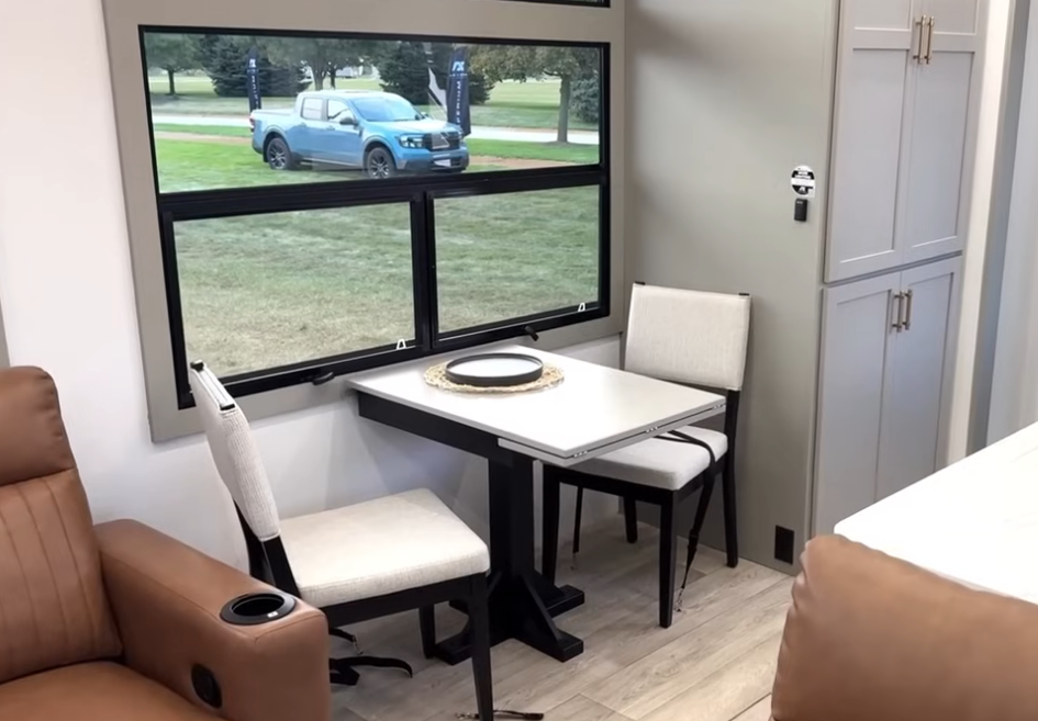Compact dining nook inside the 2026 Axiom Imperium I3800 featuring a modern table with two cushioned chairs beside a large panoramic window, offering a bright and inviting dining space with scenic outdoor views.