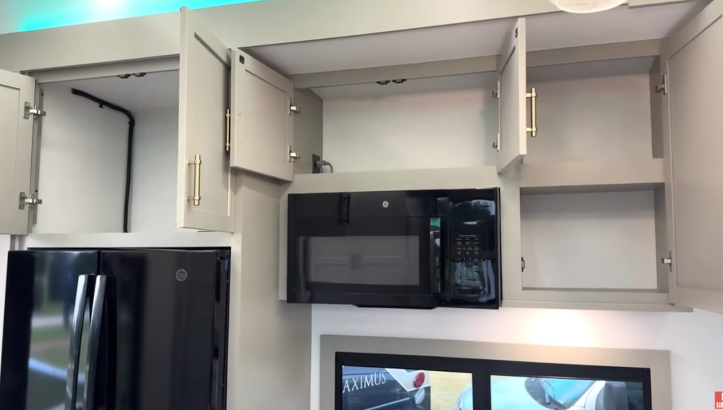 Open kitchen cabinets inside the 2026 Axiom Imperium I3800 fifth wheel showing generous overhead storage, a GE refrigerator, built-in microwave, and modern cabinetry with gold hardware and LED accent lighting.