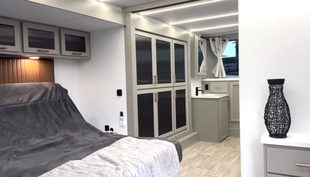 Bedroom suite inside the 2026 Axiom Imperium I3800 fifth wheel featuring a large bed with overhead storage, built-in wardrobe cabinets, vanity with sink, and modern LED lighting for a luxurious residential feel.