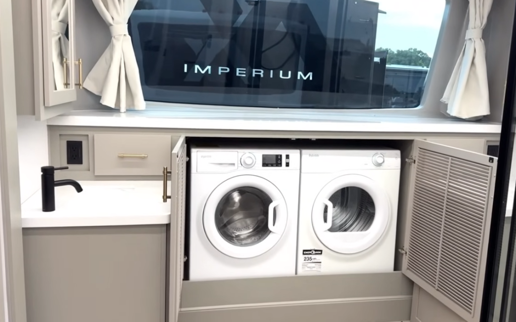 Bedroom laundry area inside the 2026 Axiom Imperium I3800 featuring a built-in washer and dryer, vanity with black faucet, large window with curtains, and stylish cabinetry for a residential RV experience.