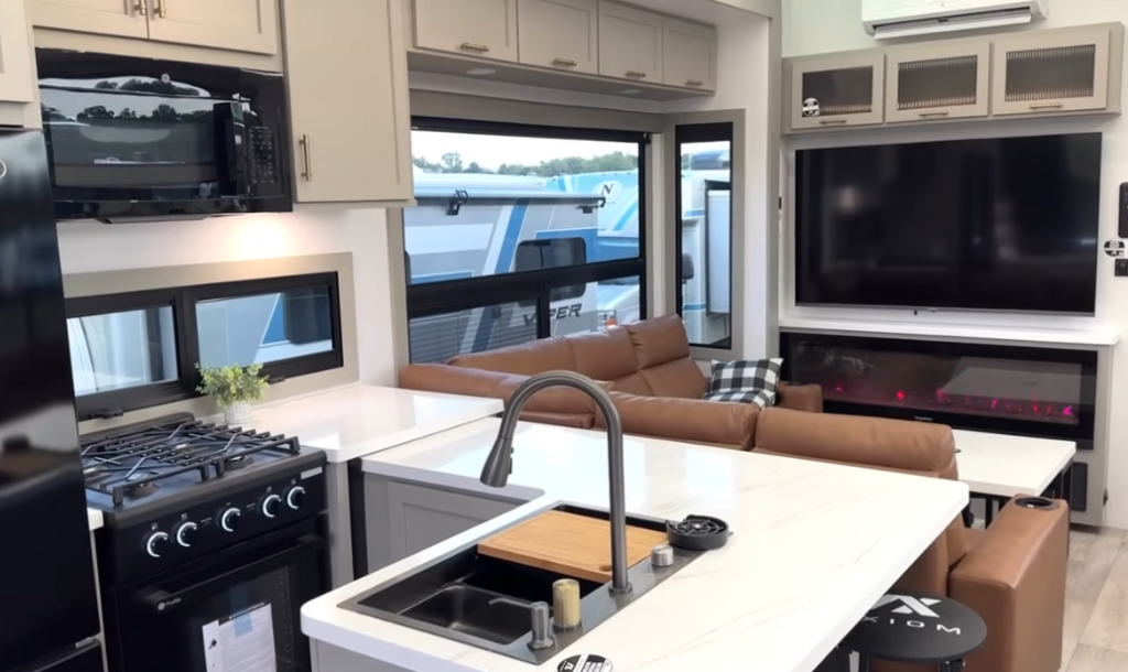 Luxury kitchen and living space inside the 2026 Axiom Imperium I3800 featuring a center island with sink, modern gas range, GE refrigerator, large TV with fireplace, and a brown leather sectional for an upscale residential feel.
