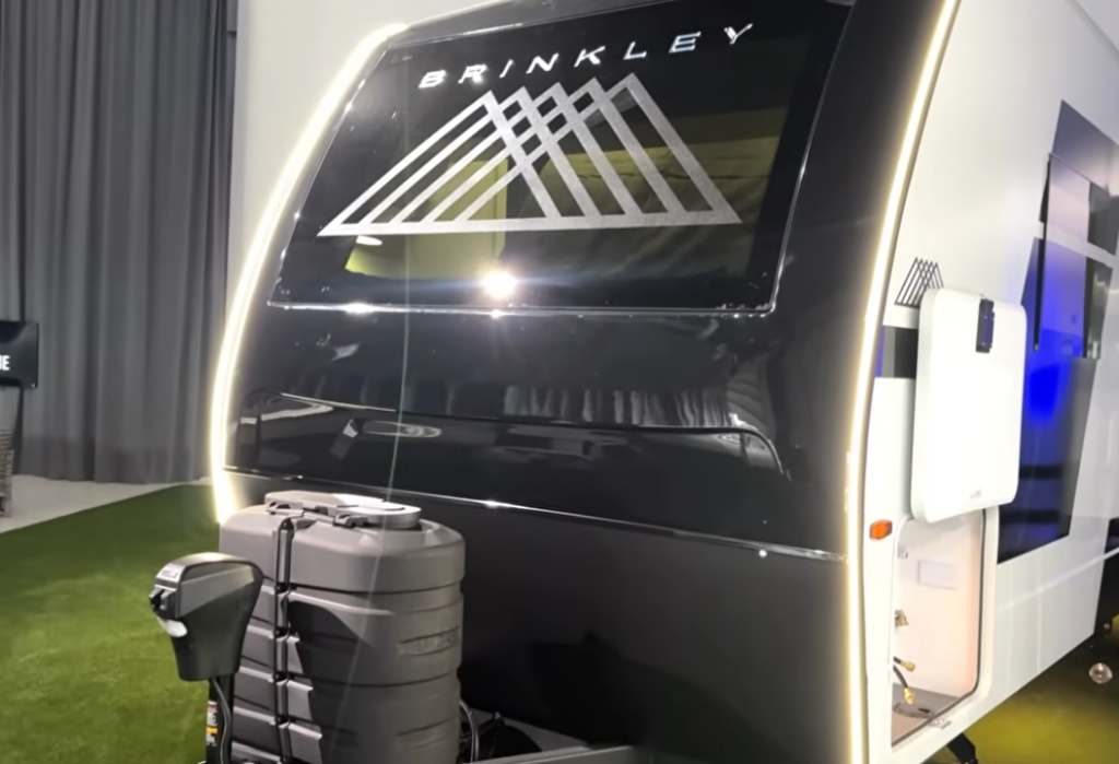 Front cap of the Brinkley Model I 280 showcasing its sleek automotive design with integrated LED accent lighting and a large dual-pane windshield. The bold Brinkley logo and aerodynamic shape emphasize the trailer’s premium construction and modern styling, with dual propane tanks and power jack visible at the hitch.