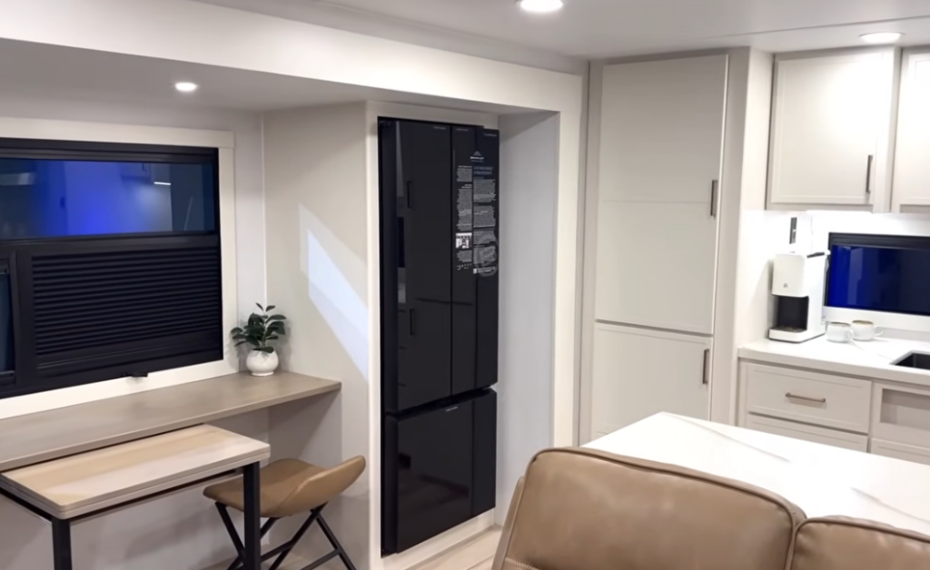 Interior view of the Brinkley Model I 280 showcasing the multifunction dinette and modern kitchen layout. The image features a fold-out dining and workspace area beside a sleek black 12V refrigerator, with white cabinetry, solid-surface countertops, and a built-in coffee maker under bright LED lighting—highlighting the RV’s open, residential design.