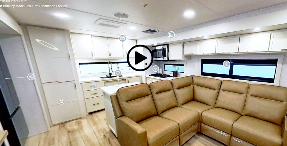 Interior view of the Brinkley Model I 280 showcasing its open-concept living and kitchen area with tan leather quad-recliner seating, solid-surface island, and modern cabinetry. The space features a stainless steel microwave, large windows, and residential lighting, highlighting the RV’s luxurious and spacious design.
