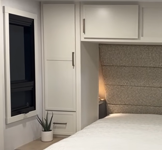 “Brinkley Model Ix 20X bedroom corner with side wardrobe, window, nightstand, and soft lighting beside the padded headboard for a cozy modern RV design.