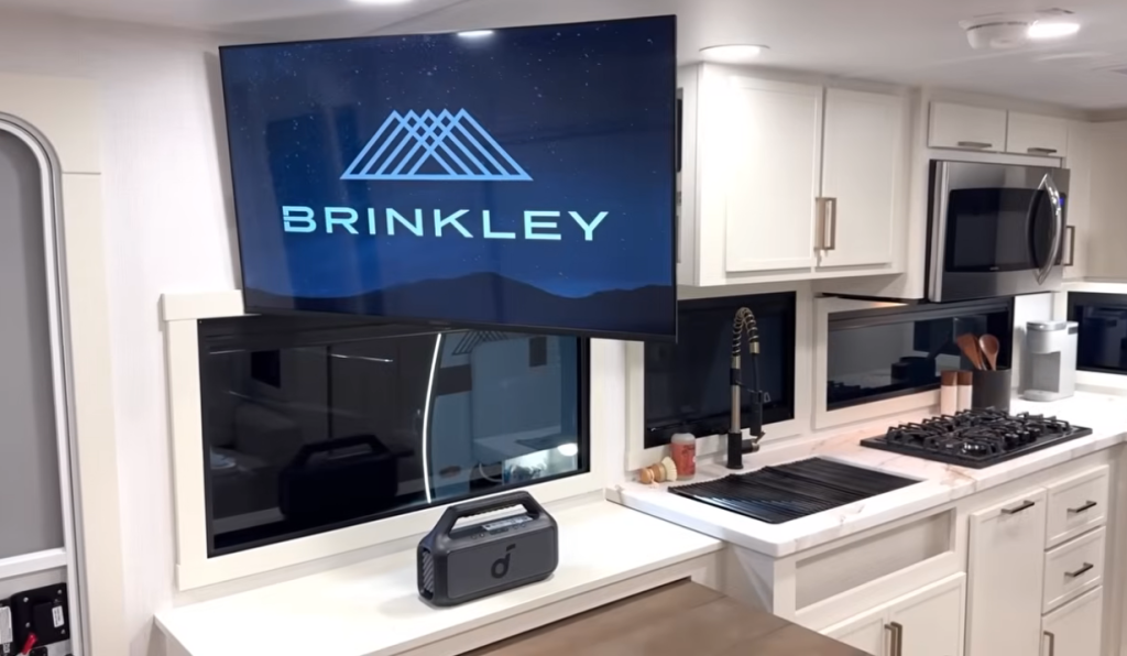 Brinkley Model Ix 20X kitchen with marble-look counters, three-burner stove, stainless microwave, farmhouse faucet, and swivel TV displaying the Brinkley logo above wide panoramic windows.