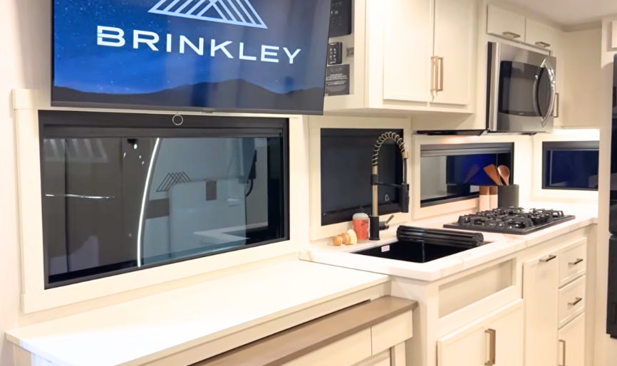 Brinkley Model Ix 20X kitchen with marble-look counters, farmhouse sink, high-arc faucet, stainless microwave, and swivel TV above panoramic windows in a luxury travel trailer.