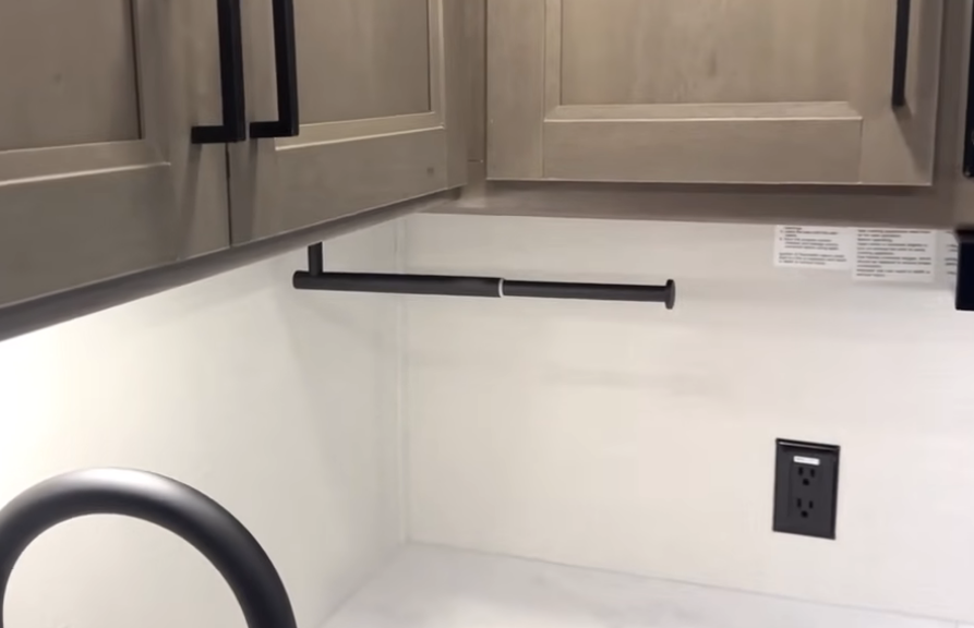 Grand Design Transcend Xplor 22RBX kitchen detail showing black hanging rail beneath upper cabinets, matte black faucet, and outlet for appliance convenience.