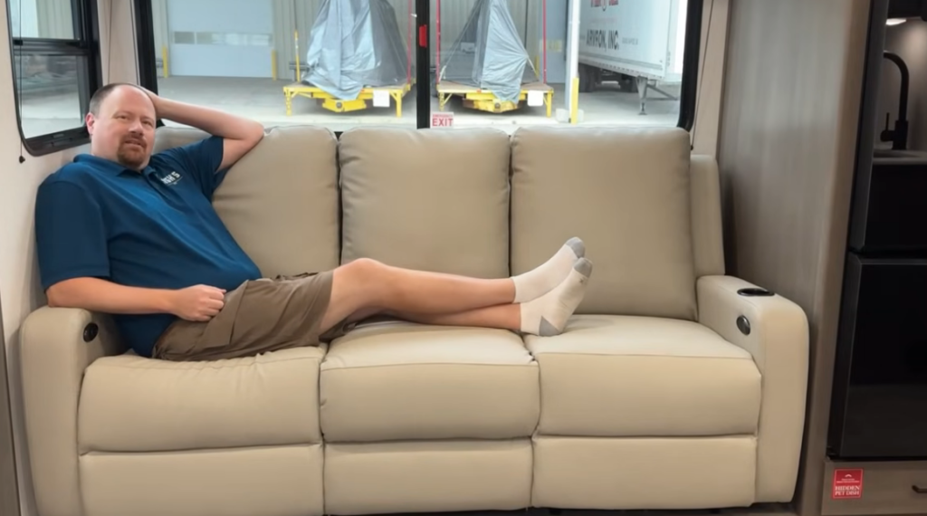 Man relaxing on tan reclining sofa inside the Grand Design Transcend Xplor RV, demonstrating spacious and comfortable interior seating design.