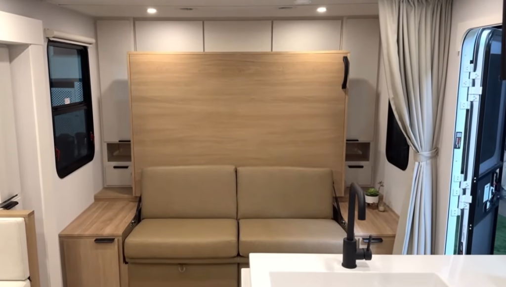 Winnebago Thrive RV living area featuring a convertible Murphy bed and sofa setup with light wood cabinetry, side tables, and natural light from surrounding windows—showcasing a modern, space-saving design ideal for couples or small families.