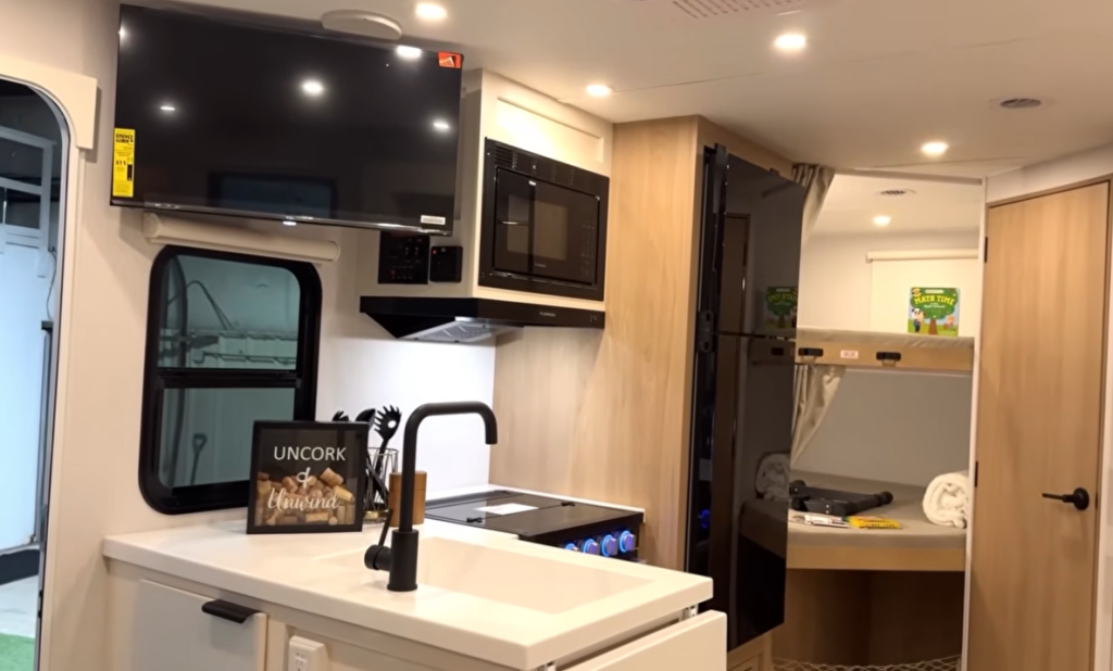 Winnebago Thrive 26T travel trailer kitchen and living space with modern light wood cabinetry, large flat-screen TV, built-in microwave, black faucet sink, and bunk beds in the rear—showcasing a bright, functional layout ideal for family RV travel.