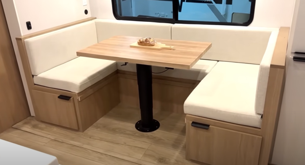 Winnebago Thrive RV dinette area with light wood cabinetry, beige cushioned seating, under-bench storage drawers, and a natural wood tabletop beneath a large window—designed for dining, lounging, or converting into an extra sleeping space.
