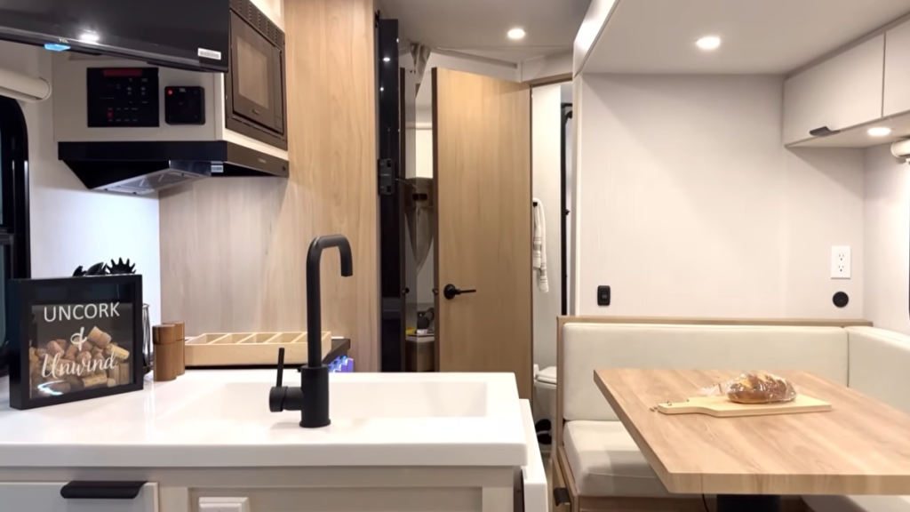 Interior of the Winnebago Thrive travel trailer showcasing the modern kitchen and dining area, featuring a white farmhouse sink with matte black faucet, overhead microwave, wood-grain cabinetry, and a cozy U-shaped dinette with natural light and soft LED ceiling lighting.