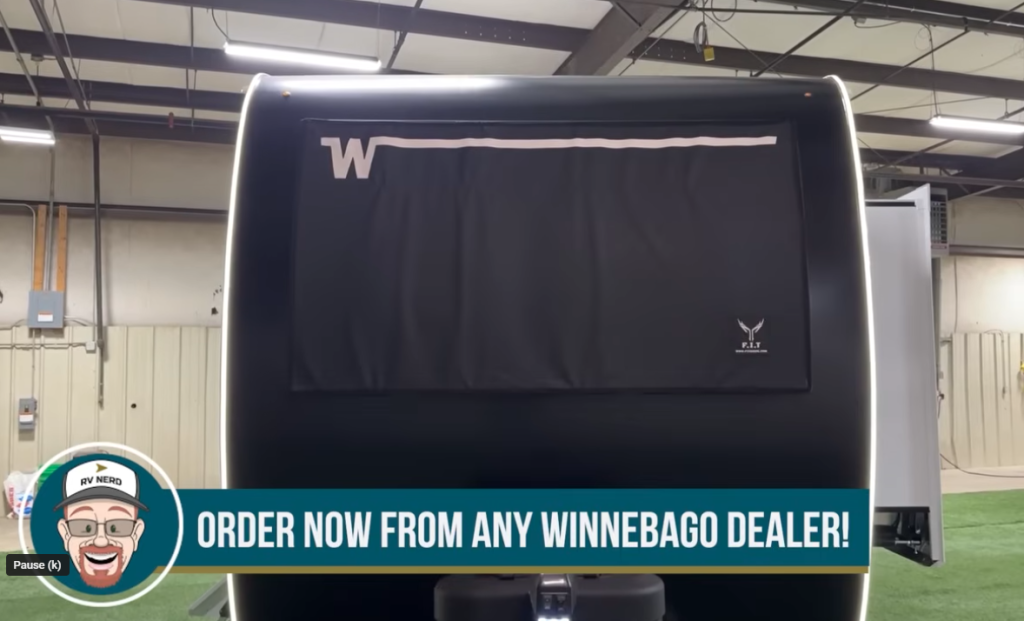 Front view of a Winnebago Thrive travel trailer with a black protective cover over the panoramic window, LED accent lighting along the edges, and text overlay reading “Order Now from Any Winnebago Dealer,” displayed inside a showroom setting.